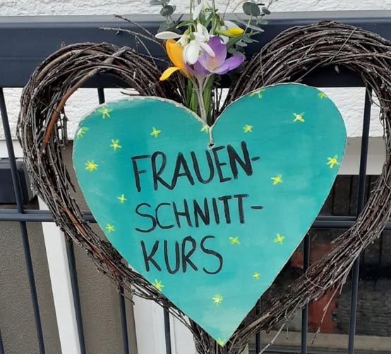 Herzförmige Dekoration aus Zweigen mit grünem Schild und der Aufschrift „Frauenschnittkurs“, geschmückt mit Blumen und aufgehängt an einem Zaun.